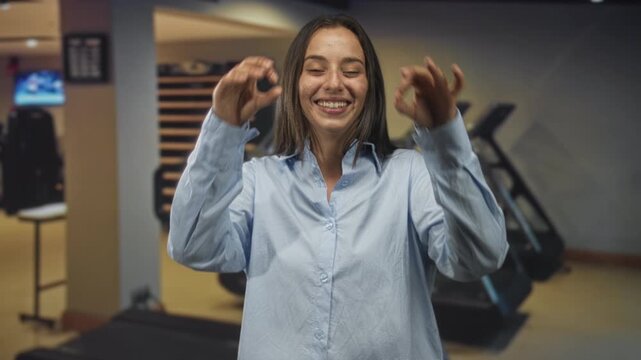 Woman forming binocular circles with hands over eyes in gym building, smiling broadly in a loose button shirt while treadmills line the room; joyful moment.