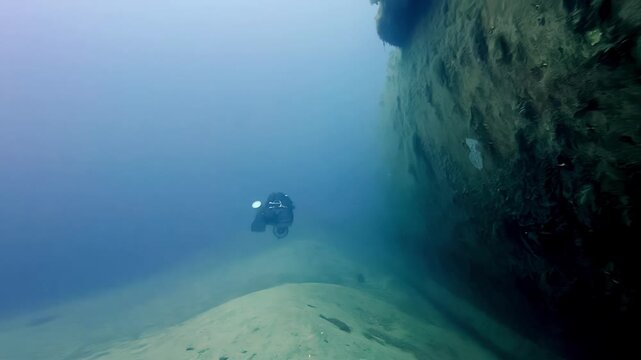 Scuba diver exploring a massive rusted shipwreck hull resting on a sandy seabed during a clear summer day in the deep blue Mediterranean Sea captured from a low wide angle showing scale and mystery