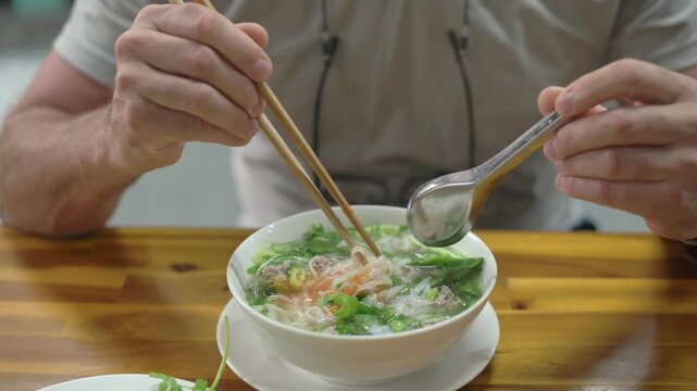 Caucasian Critic Examines Pho Ingredients Carefully. Person Uses Chopsticks And Spoon To Taste Vietnamese Soup. Food Enthusiast Carefully Inspects Aromatic Beef And Noodles At Busy Cafe