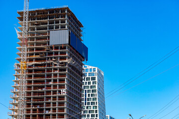 Fototapeta premium High-rise under construction against clear blue sky. Steel frame rises with scaffolding and cranes. Modern building beside finished structure. Urban development in progress, bold and active