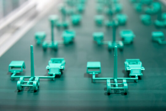 A precision close-up shot captures numerous small, teal-colored plastic components, possibly parts for toy vehicles or prototypes, moving along a dark green conveyor belt