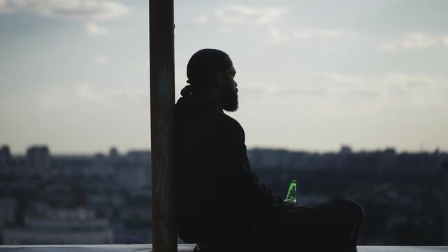 Young man in a durag sits on rooftop edge and drinks beer, lost in thoughts