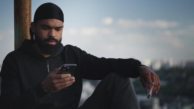 Young man selling pot online, texting on phone on city rooftop, illegal business