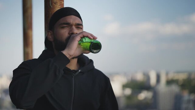 Young Black man in a hoodie drinks beer on city rooftop, thinking about his life