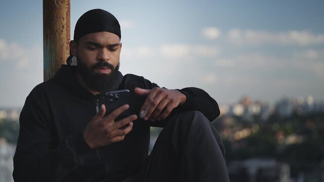 African American man in durag texting on phone on rooftop, holding pot in hand