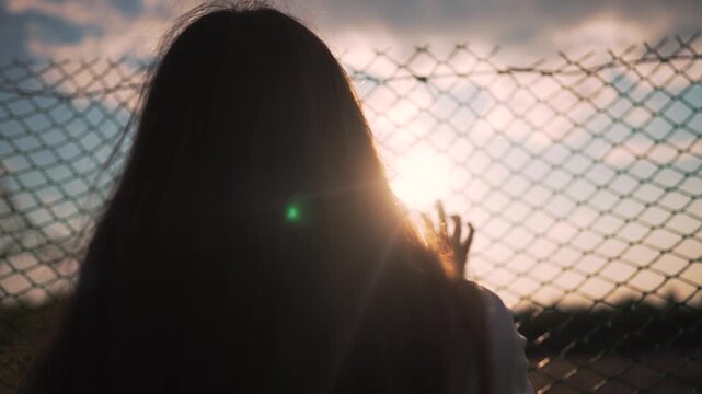 Woman touching chainlink fence at sunset with hand on wire mesh. Girl places hand on fence barrier outdoors. Silhouette shows woman at chainlink fence. Girl touching wire fence at sunset sky.