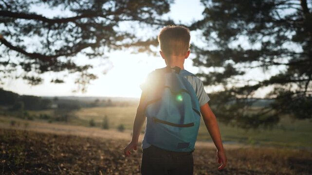Boy with backpack walks outdoors at sunset looking at forest landscape. Child explores nature with backpack during evening. Boy hiking outdoors at sunset. Kid with backpack walks in countryside.