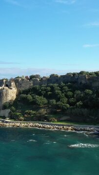 Scenic view of an old stone fortress surrounded by lush greenery along the coastline in Koroni, Greece, under a clear blue sky.