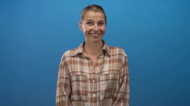 Woman smiling with eyes closed wearing a brown plaid shirt in a studio with a blue backdrop; contentment.