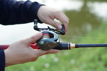 Close-up of hands adjusting a baitcasting fishing reel on a rod near calm water, preparing for angling outdoors, showcasing fishing gear detail and recreational activity in nature.