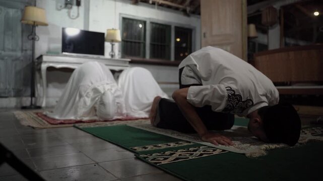 Asian father leading sujud with daughters, family performing prostration together on prayer mats inside rustic home, synchronized