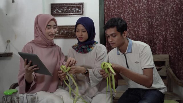 Asian family weaving palm craft in cozy living room, woman in pink hijab holds tablet while younger woman watches and man
