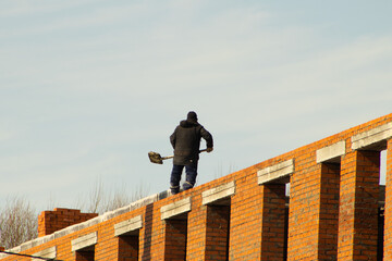 A construction worker dressed in dark winter clothing stands on the narrow edge of a high brick wall under a clear sky. The worker is holding a shovel, engaged in manual labor © Andrey