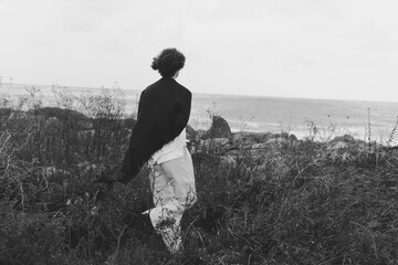 Naklejka premium Silhouette of a woman walking through meadow near coast in grayscale, wind in hair, tranquil and solitary journey amid sea spray and wildflowers