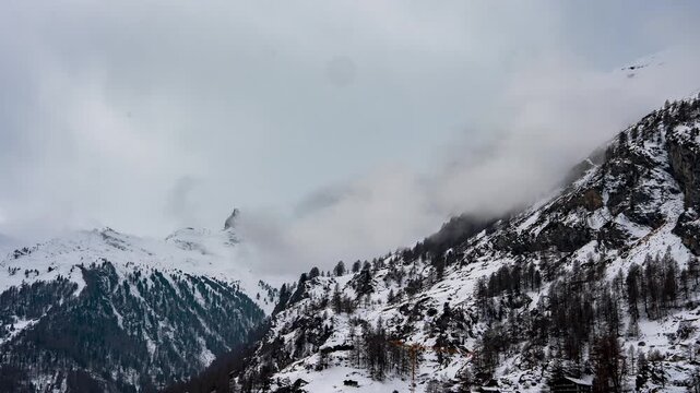 Timelapse shows the Matterhorn above Zermatt as clouds and fog sweep ridgelines, veiling the summit. Snow covers rocky faces and dark conifer bands near chalets.