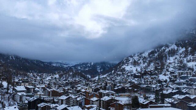 Wide timelapse in Zermatt, Switzerland, shows chalets and a church as fog and low clouds sweep by, the Matterhorn is concealed, snow flurries fall, cool winter light.