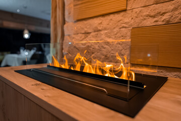 Fototapeta premium Close up interior shows a modern linear fireplace behind glass in a wood and stone console, warm light on textured stone, with a dim dining area in Zermatt, Switzerland.