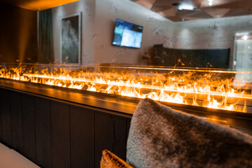 Fototapeta premium A linear fireplace flickers behind glass in a modern alpine lounge in Zermatt, Switzerland. Warm amber light plays on cushions, dark wood, and a wall mounted TV.