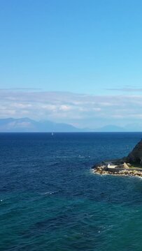 Clear blue sea with a rocky coastline under a bright sky in Koroni, Greece. A distant sailboat is visible on the horizon, creating a serene Mediterranean scene.