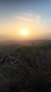 Golden sun setting over a hazy mountain landscape in the Canary Islands, with arid vegetation and rocks in the foreground creating a serene and dusty atmosphere during a beautiful summer evening