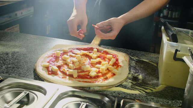 Chef places salami pieces by hand building topping pattern across crust. Pizza base shows tomato sauce and cheese cubes on peel near prep station