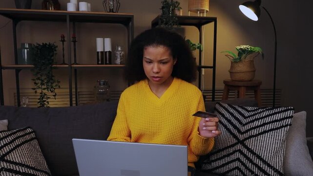 Unhappy confused African American young woman has problem with online shopping blocked credit bank card laptop error payment mistake security number angry sad girl female with computer at evening home
