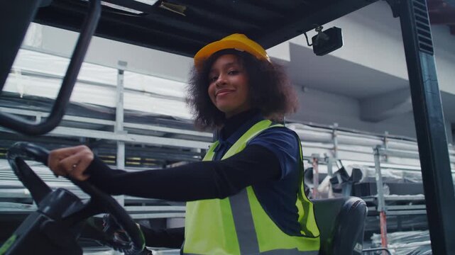 Portrait of a smiling female worker wearing protective gear, a yellow hard hat, and a safety vest, driving a forklift truck in a warehouse.
