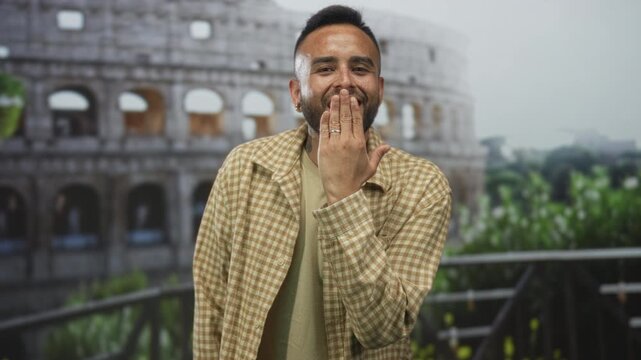 Man in front of coliseum building blows a kiss with hand to mouth while smiling at camera; friendly greeting joy.