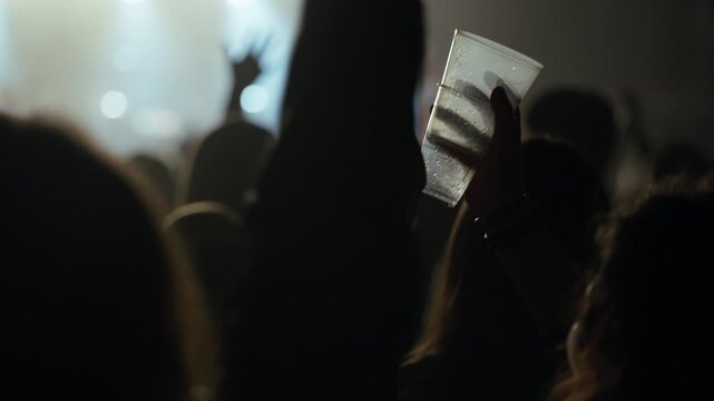 Silhouetted crowd dances enthusiastically at a live music concert or club. Flashing stage lights illuminate the smoky atmosphere as one person holds a plastic cup, swaying to the music