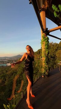 Elegant woman in black silk dress and orange heels enjoying sunset at beach club in Phuket, Thailand, overlooking Andaman Sea and Karon Beach. Luxury lifestyle, tropical evening atmosphere.
