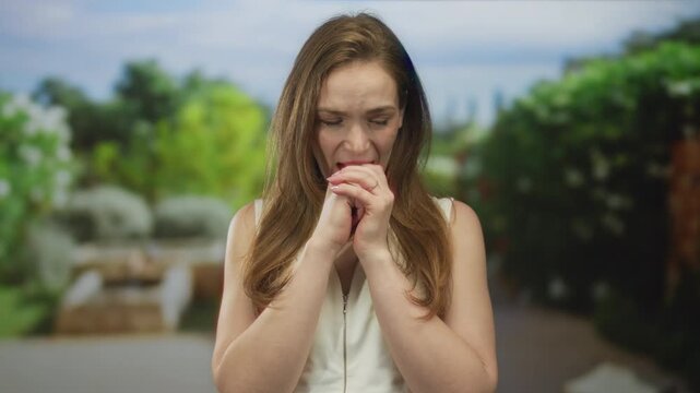 Woman biting knuckles with pained expression under leafy canopy in forest near stone bench; anxiety.