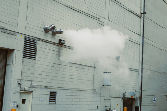 Smoke billows from vents on a factory wall during daytime in an industrial area