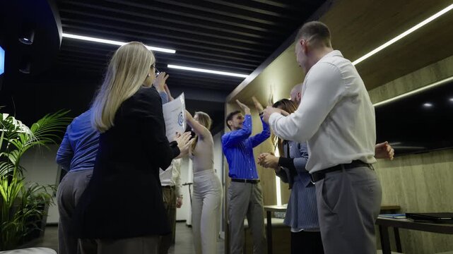 A diverse group of business professionals celebrates a shared success with high-fives and joyful embraces. Happy colleagues show teamwork and achievement in an office setting.