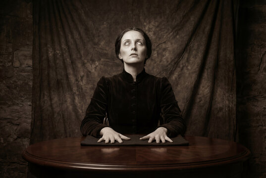 Vintage sepia portrait of an eerie pale medium with blank white eyes performing a spiritual seance at a table. Dark occult horror concept.