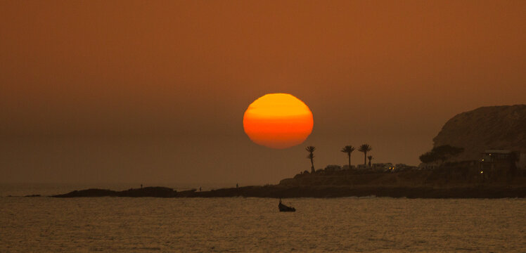 The setting sun descends over the Atlantic Ocean towards the cliffs as people play in the sea and on the beach at the end of another hot day in Taghazout, Agadir, Morocco