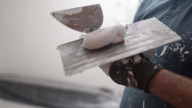 Close up video of a construction worker holding a trowel and applying putty or plaster on an interior wall during renovation work.