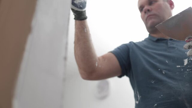 Low angle video of a construction worker using a trowel to smooth plaster or putty on an interior wall during home renovation.