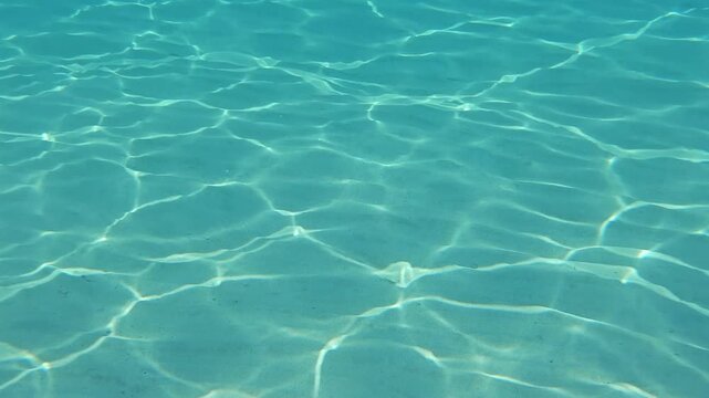 Underwater video of a sandy seabed in clear shallow sea water with natural sunlight patterns and gentle movement below the surface.