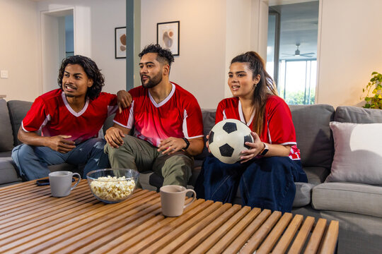 Asian friends in red jerseys watching match at home on sofa, woman holding soccer ball, popcorn