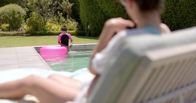 African American woman on chair watching female child easing pink tube, entering pool with goggles