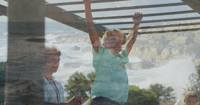 Boy gripping bar, swinging across monkey bars, coach recording sports skills, faint coastal overlay