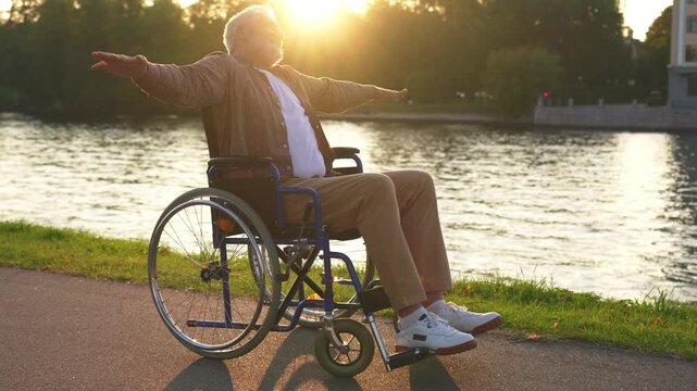 Happy senior man in wheelchair raised hands enjoying freedom walking on road in city park outdoor. Old happy man in chair for people with disability has a hope. Disability disease rehabilitation