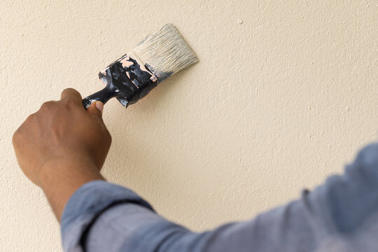 African American man painting textured beige wall at home in blue gray shirt with wide paintbrush