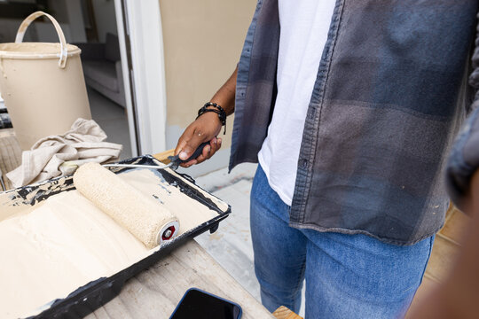 African American man prepping paint roller with beige tray and phone near open doorway, copy space