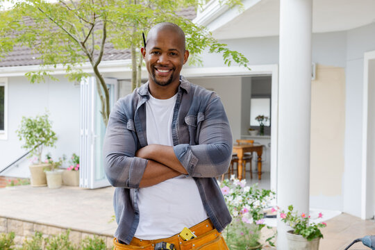 Smiling handyman standing with arms crossed on front porch, wearing grey plaid and yellow tool belt
