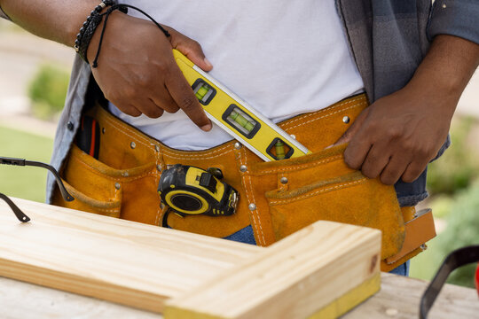 African American man wearing denim shirt holding yellow spirit level on tool belt at backyard
