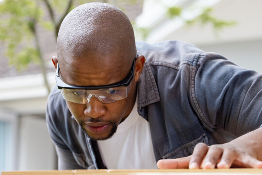 African American man leaning in, inspecting wooden plank on home porch while wearing safety goggles