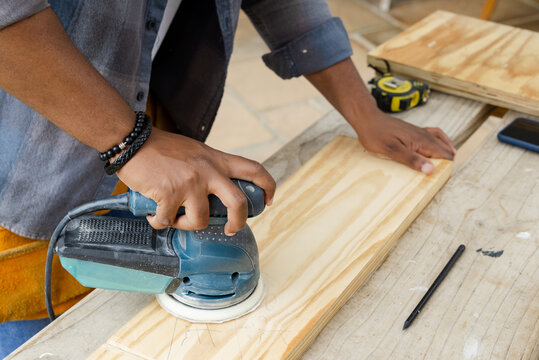Man sanding pine plank with orbital sander at workbench wearing denim shirt and yellow apron