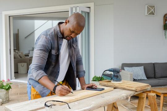 Male woodworker leaning over workbench, marking plank using smartphone reference, prepping cuts
