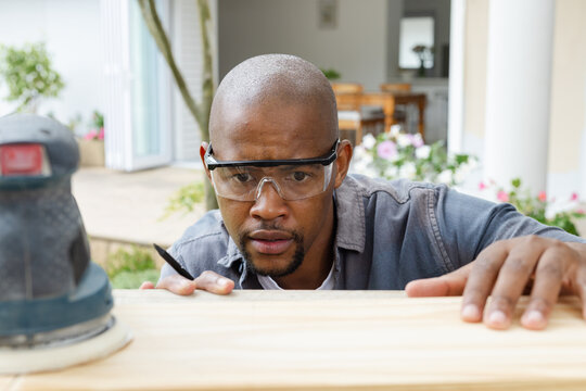 Checking man wearing grey shirt goggles leaning over wood plank porch pencil behind ear sander near